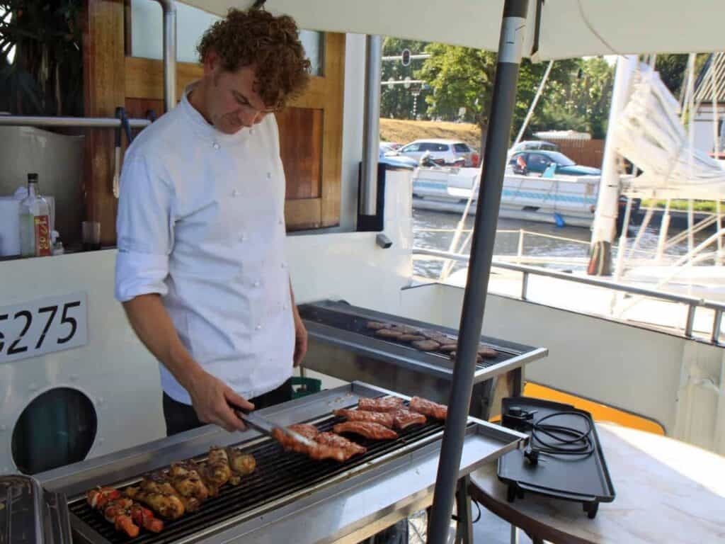 Chef bereidt vlees op barbecue op een partyboot, genietend van de zon en het uitzicht.